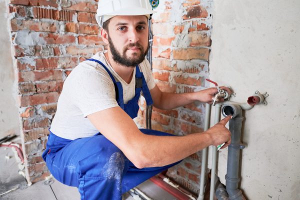 Handsome male worker in safety helmet looking at camera while applying silicone grease on pipe. Man plumber in work overalls lubricating pipe while installing water system in apartment.