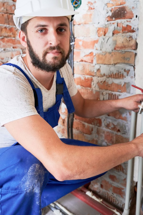 Handsome male worker in safety helmet looking at camera while applying silicone grease on pipe. Man plumber in work overalls lubricating pipe while installing water system in apartment.