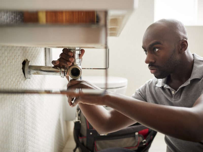 Young black male plumber sitting on the floor fixing a bathroom sink, close up